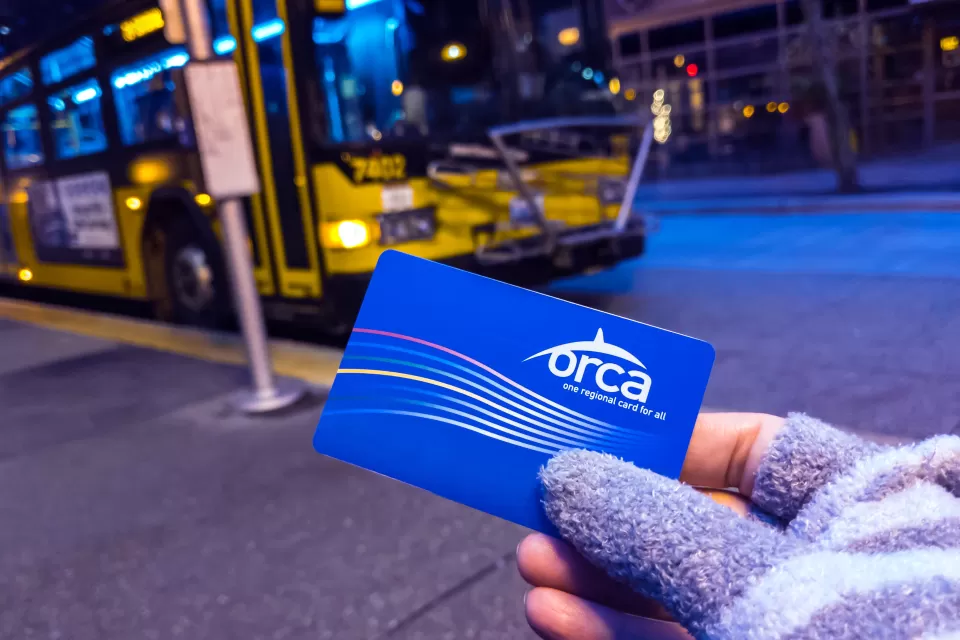 Close-up of gloved hand holding blue Orca transit card with blue and yellow bus in background